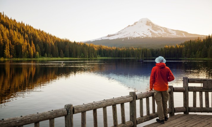 Man in a red jacket standing on a platform near a lake looking at Mt Hood