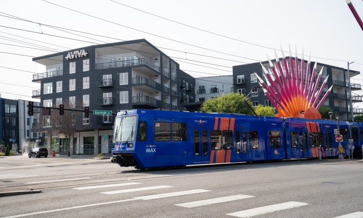 A blue Trimet MAX train glides past a dense housing development in Gresham