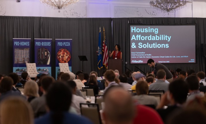 A woman in a red dress standing on a stage behind a lectern, in front of a large crowd. On the stage with her are some banners that say, "Pro-housing," "Pro-riders," and "Pro-pizza." There is also a presentation projected on a screen that says, "Housing Affordability and Solutions."