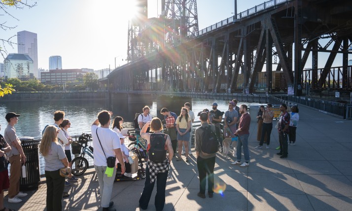 A group of people gathered on a paved path along a river, next to a bridge, and across the water from skyscrapers.