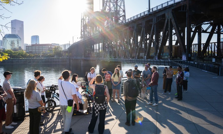 Group of people gathered on a paved, multi-use path along a river, with a large bridge and skyscrapers in the background