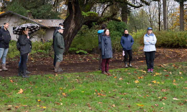 A group of people standing on the edge of an agricultural field, with a moss-laden tree behind them