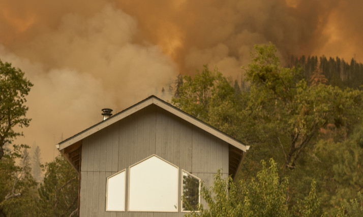 A cloud of smoke and an orange sky rise above a home in a forested area