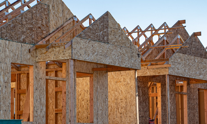 Housing being built, with frames and plywood walls up. On the right side, a man on a ladder is hammering on one of the walls