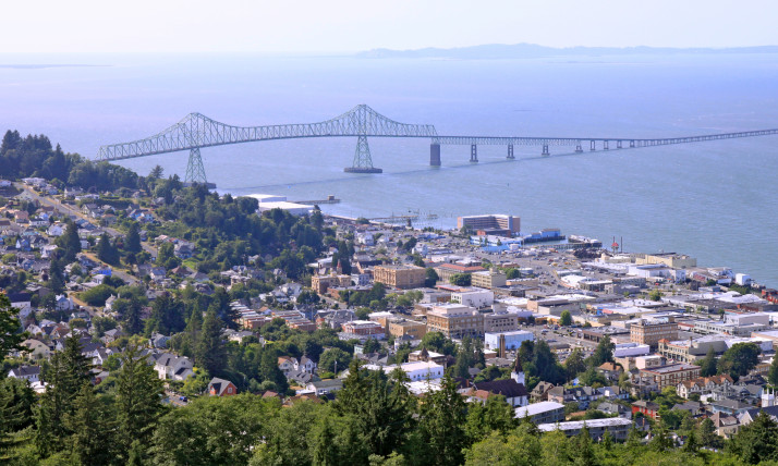Aerial view of Astoria and the Astoria-Megler Bridge across the wide, blue Columbia River. Astoria itself is tucked between a forested hillside and the river's shore. The densest development is near the water, on flatter ground. The forested hillsides have a smattering of homes among the trees.