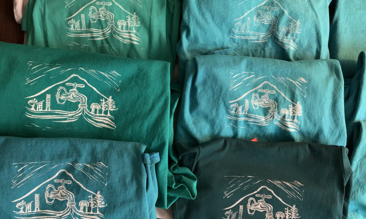 Green, blue, and teal shirts with a white print on them. The design has a faucet of flowing water in the middle, which forms a river that is flowing past a tiny city on one side, and a rural location on the other side. All of this is encircled by an outline of a pointed peak (Mt. Hood).