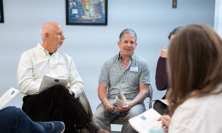Five people sit on chairs arranged in a circle in an indoor setting, holding papers and wearing nametags. They appear to be talking to one another.