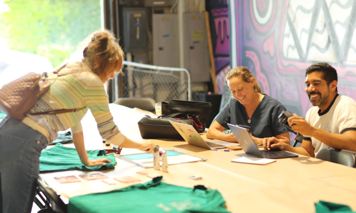 A group of people gathered around a large table in a space filled with colorful murals. One person is leaning over the table to decorate a teal shirt. Two others are smiling while working at a laptop.