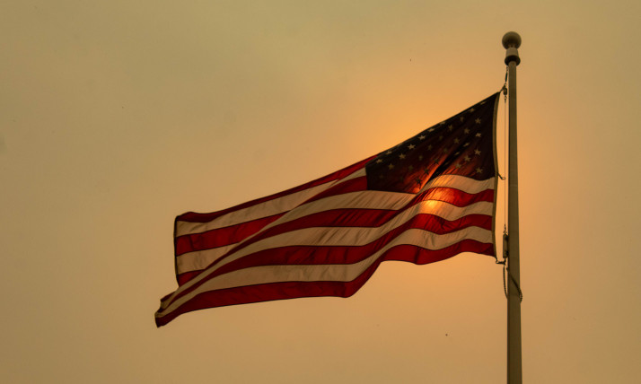 An American flag backlit by an orange sun, against a brown-orange sky hazy with wildfire smoke