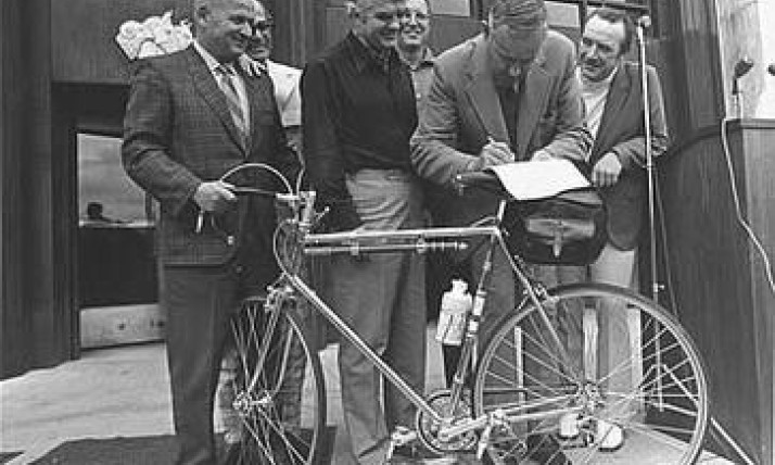 A group of men gathered around a bike; one man, Tom McCall, is signing a paper on the bike seat.