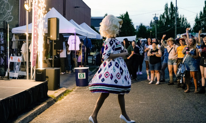 A tall drag queen in a large, platinum blond wig; a floral dress with a wide, knee-length skirt; and white high heels performs in front of a cheering crowd at a street fair with booths in an outdoor, urban setting