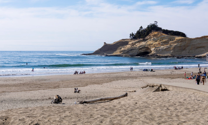 A sandy beach with blue waters and a prominent sandstone formation jutting out to sea. There are many people scattered along the beach, walking, running, and playing.