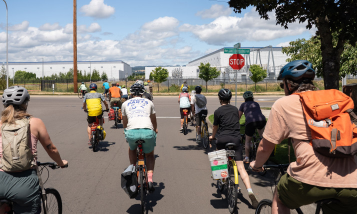 A group of people on bikes in front of a few large, windowless, white buildings (data centers)