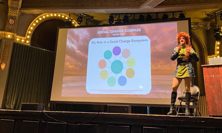 A drag queen in a green mini dress with knee-high boots and a long, red wig, standing in front of a presentation screen that says, "Social change compass."