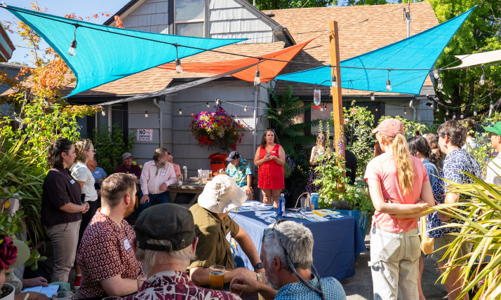 A group of people in a colorful, outdoor patio space. Everyone is looking towards a woman in a red dress, who is speaking.