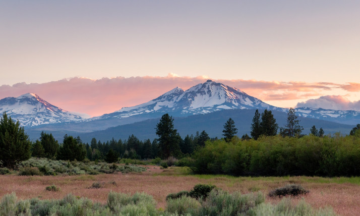 Three pointed, glaciated mountains rise above a flat plain with grass, sagebrush, and some Ponderosa pines