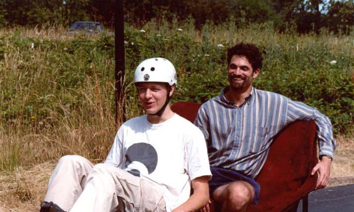 A pedal-cab, with a young man in a white shirt and white helmet pedaling in front, and a young man in a blue striped shirt sitting on a velvet seat in back