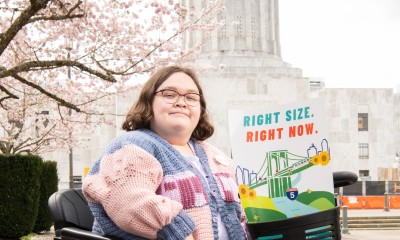 Cassie sits on a mobility device facing the camera, with a poster that shows a bridge and says "Right size. Right now." She is in front of the Salem capitol.