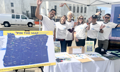 A group of 6 people smile and wave at the camera. They are standing at a booth in front of the capitol building in Salem.