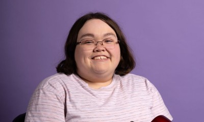 Cassie Wilson sits on an assistive mobility device against a purple background and faces the camera, smiling