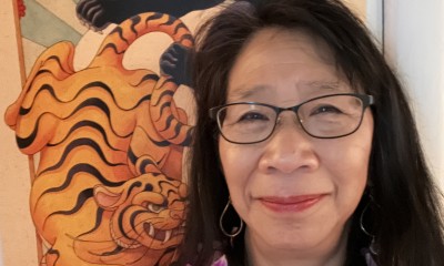 Anita faces the camera and gently smiles. She is wearing a purple lei and a piece of art hangs behind her.
