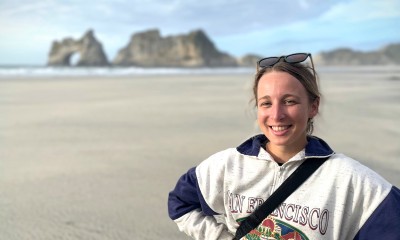 Anna stands on a beach while wearing a sweatshirt and smiles at the camera