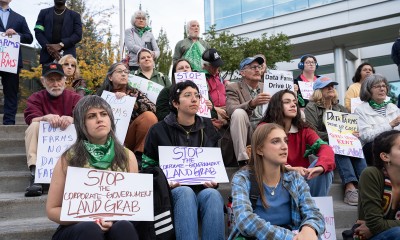 A group of people sit outside on steps and holds signs that read "stop the corporate-government land grab"