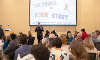 A man in a suit addressing a crowd, with a screen behind him that says, "The power of your story"
