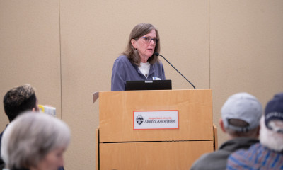 Mary Kyle McCurdy speaking at the podium, with many people looking on