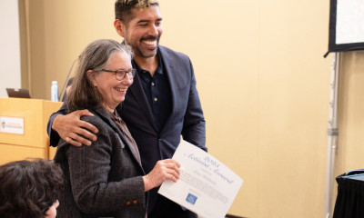 Two people smiling and sharing a side-hug; one of the people is holding a certificate