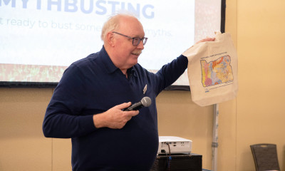 Jim Johnson holding up a tote bag with a colorful map on it; in the background is a screen that says "myth busting"