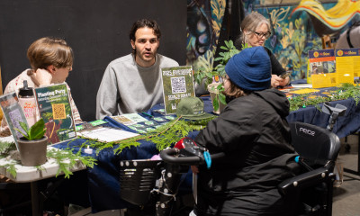 A person in a mobility scooter wearing a blue knit hat and a black coat talks to a pair of people at a booth covered in greenery