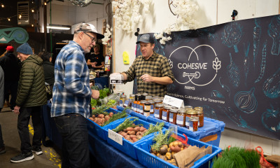 A man in a plaid shirt, buying vegetables from another man in a plaid shirt. The sign on the wall behind the booth says, "Cohesive Farms."