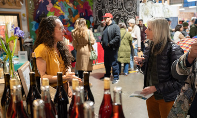 A crowded indoor market scene is framed by two people having a conversation in the foreground, and a countertop filled with bottles of wine