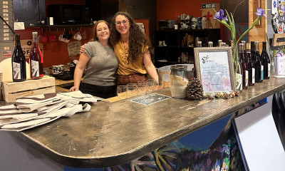 Two women smiling from behind a bar, with wine bottles and glasses on the counter