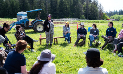 A group of people seated in chairs arranged in a circle in a grassy field near rows of agricultural crops.