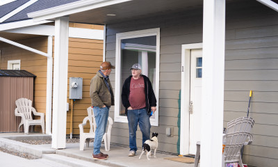 Two men and a small dog stand on the porch of a small house with white trim around the windows and door.