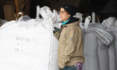 A person in a tan coat, blue jeans, and a dark beanie stands next to large (almost as tall as they are) bags of seeds in a barn