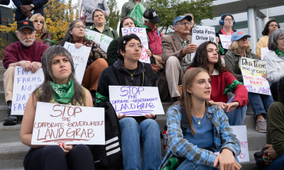 A group of people sitting on concrete steps at an outdoor rally, holding signs that say things like, "Stop the corporate-government land grab" and "food farms not data farms"