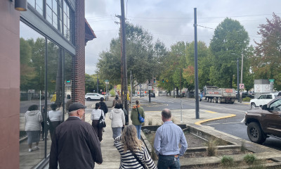 A group of people walking along a sidewalk. On the right side of them is a bioswale, and on the left is a new building with large, shiny windows.