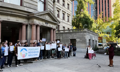 A group of people gathered around Portland City Hall holding signs, the largest of which says "Portland Neighbors Welcome"