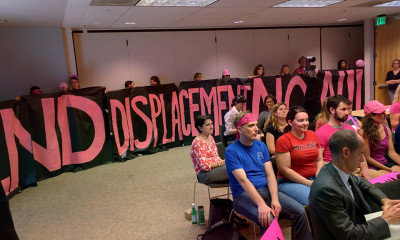 People sitting in a city council meeting with banners saying "no displacement"