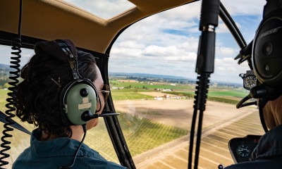 Two people with ear protection in a helicopter, looking out towards the farmland stretching below them