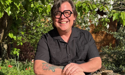 A smiling man with glasses, a navy blue shirt, and a tattoo on his arm in a garden