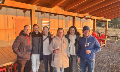 A smiling group of people under a wooden shelter. Some of the people are holding wine glasses.