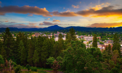 Aerial image of a city surrounded by trees and mountains, with a colorful sunset unfolding