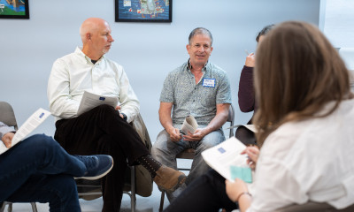 Five people sit on chairs arranged in a circle in an indoor setting, holding papers and wearing nametags. They appear to be talking to one another.