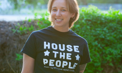 A smiling person wearing a shirt that says, "House the People--Welcome Home Coalition"