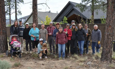 A smiling group of people, from a small child in a stroller, to older adults with gray hair, stand smiling in a group near homes and ponderosa pine trees.