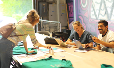 A group of people gathered around a large table in a space filled with colorful murals. One person is leaning over the table to decorate a teal shirt. Two others are smiling while working at a laptop.